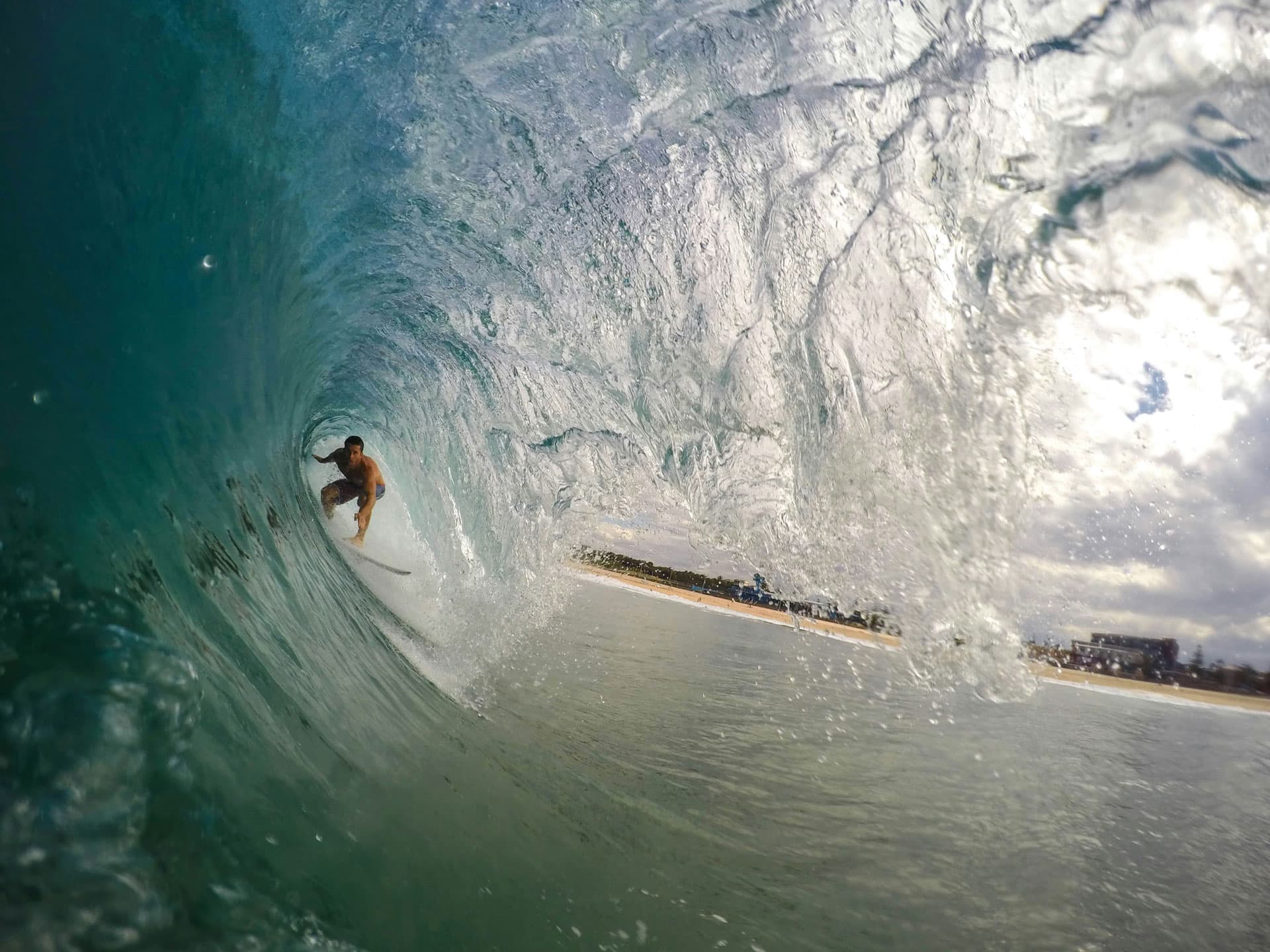 A surfer crouched inside a breaking wave, photographed from inside the barrel, with a beach and buildings behind.