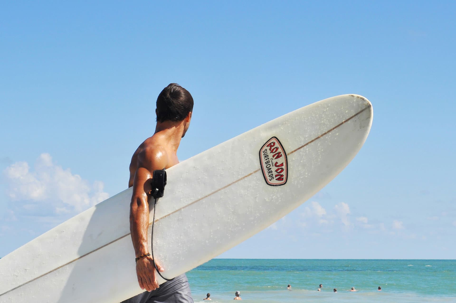A surfer seen from behind with a white longboard under one arm, the leash coiled on it and the wax and wear on the deck in view, the sea behind.