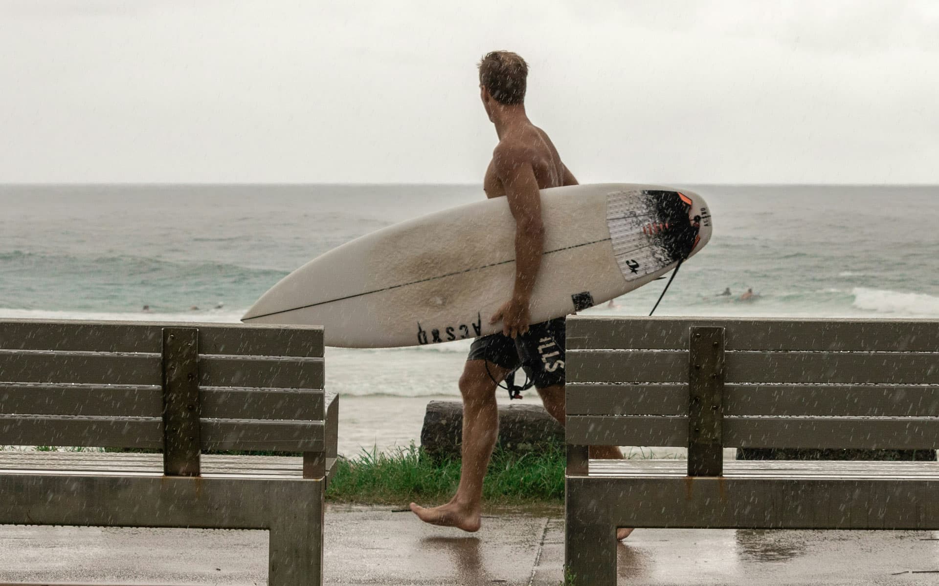 A surfer walking along a beachfront path in the rain with a well-used shortboard under his arm, the deck discoloured and a traction pad on the tail.