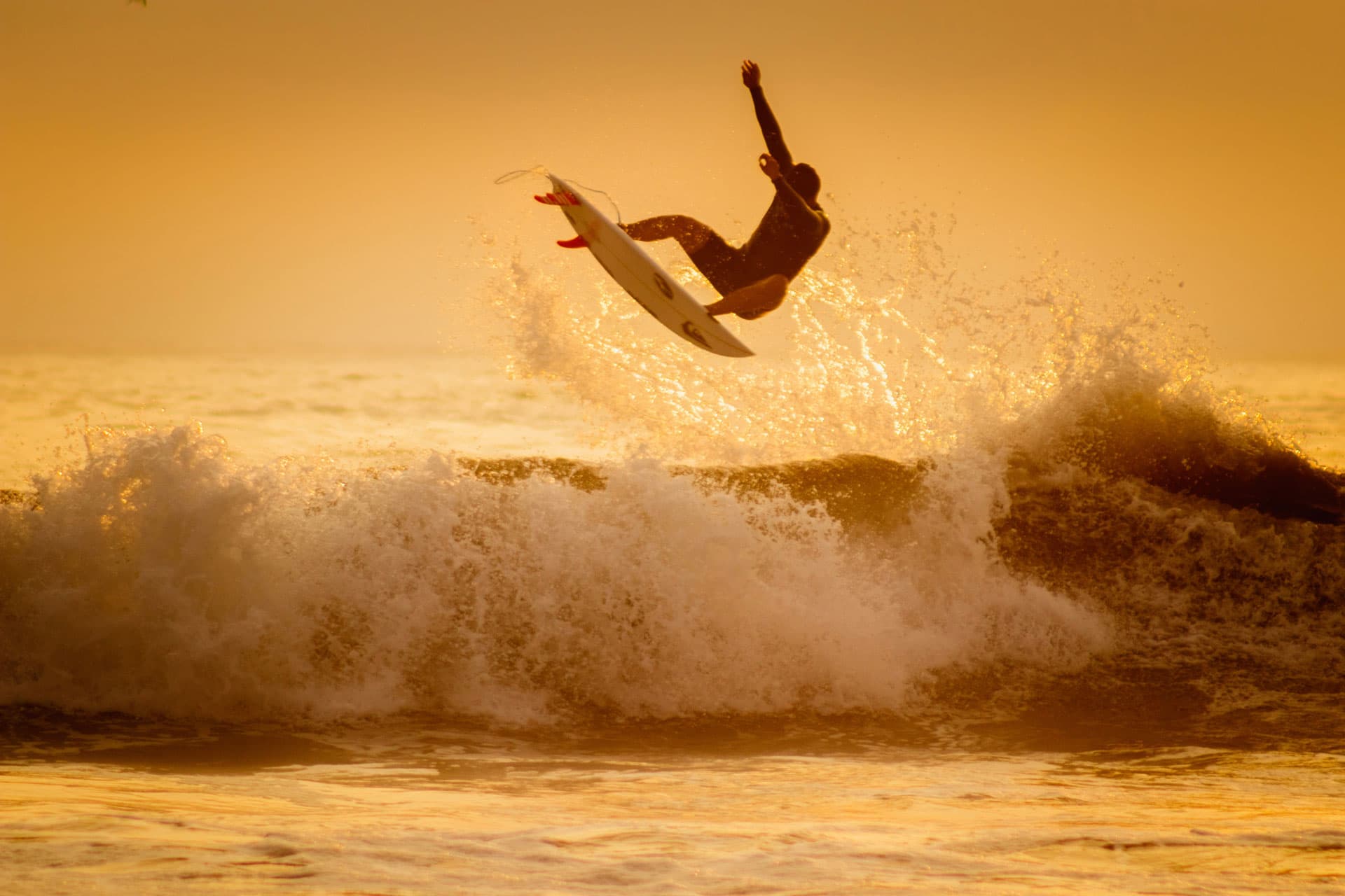 A surfer in silhouette in the air above a breaking wave at sunset, the underside of the board and its red fins turned towards the camera.