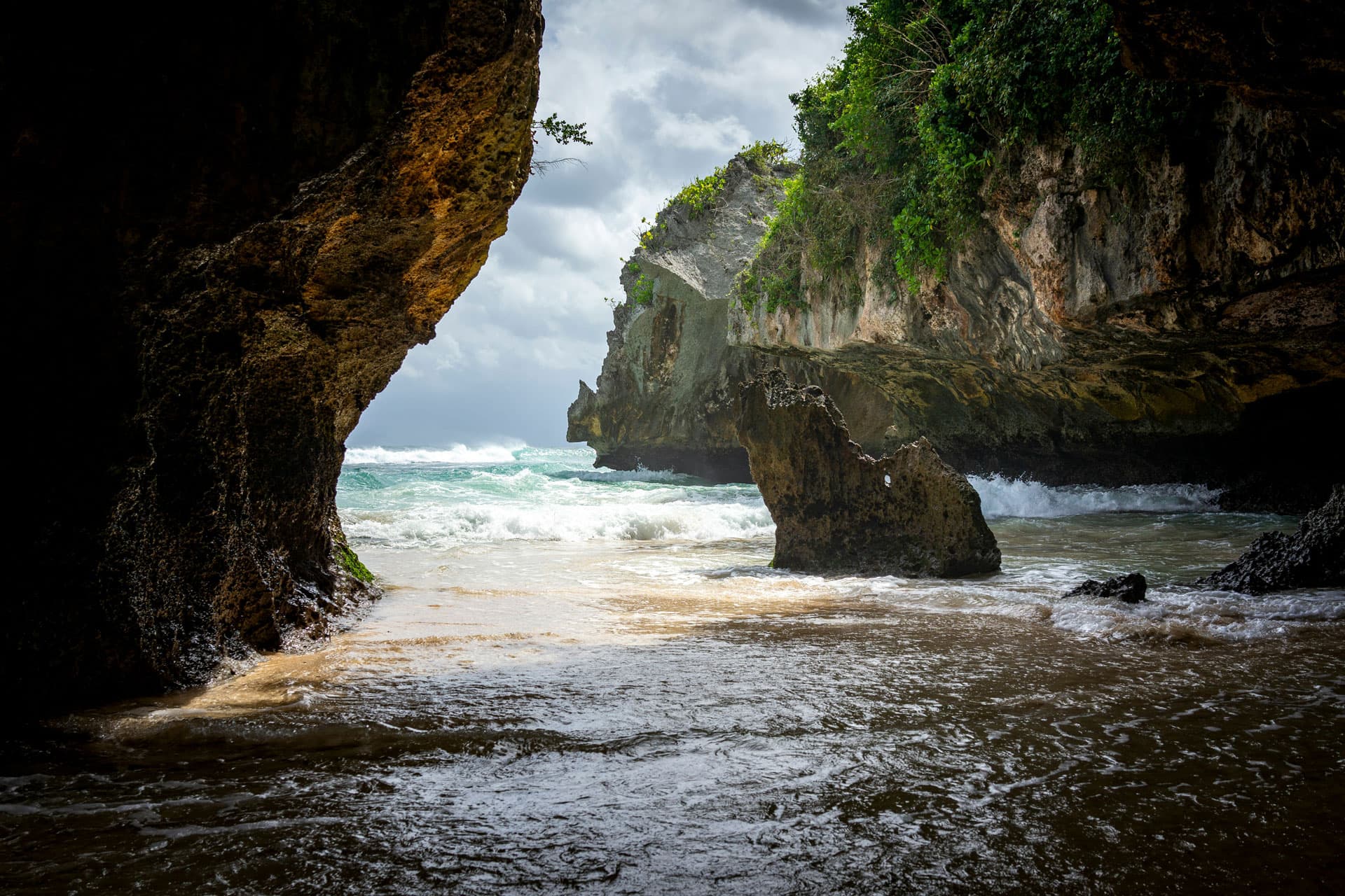 The beach below the cliffs at Uluwatu in Bali, seen from inside the cave, white water running over the sand and plants hanging off the limestone.
